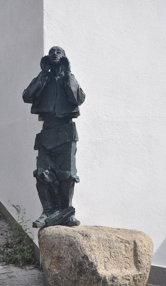 Auf einem großen Bachstein steht eine lebensgroße schwarze Männerfigur, die die Hände zum Mund hält, als würde sie schreien.