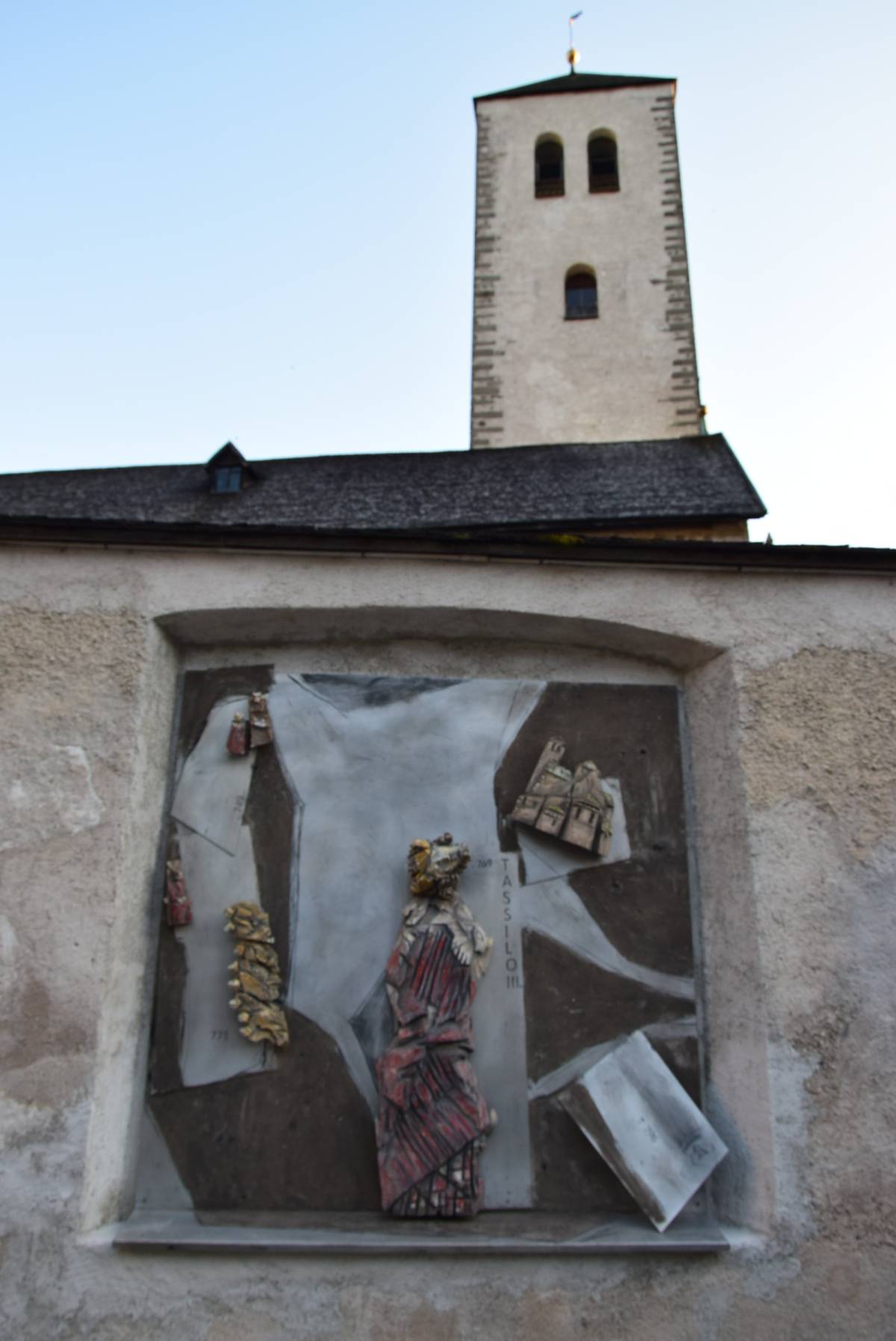 Ein Relief auf einer Aluminiumplatte, eingelassen in die Außenmauer einer Kirche. Es zeigt eine große rote Figur, umgeben von mehreren kleineren Figuren aus Holz.