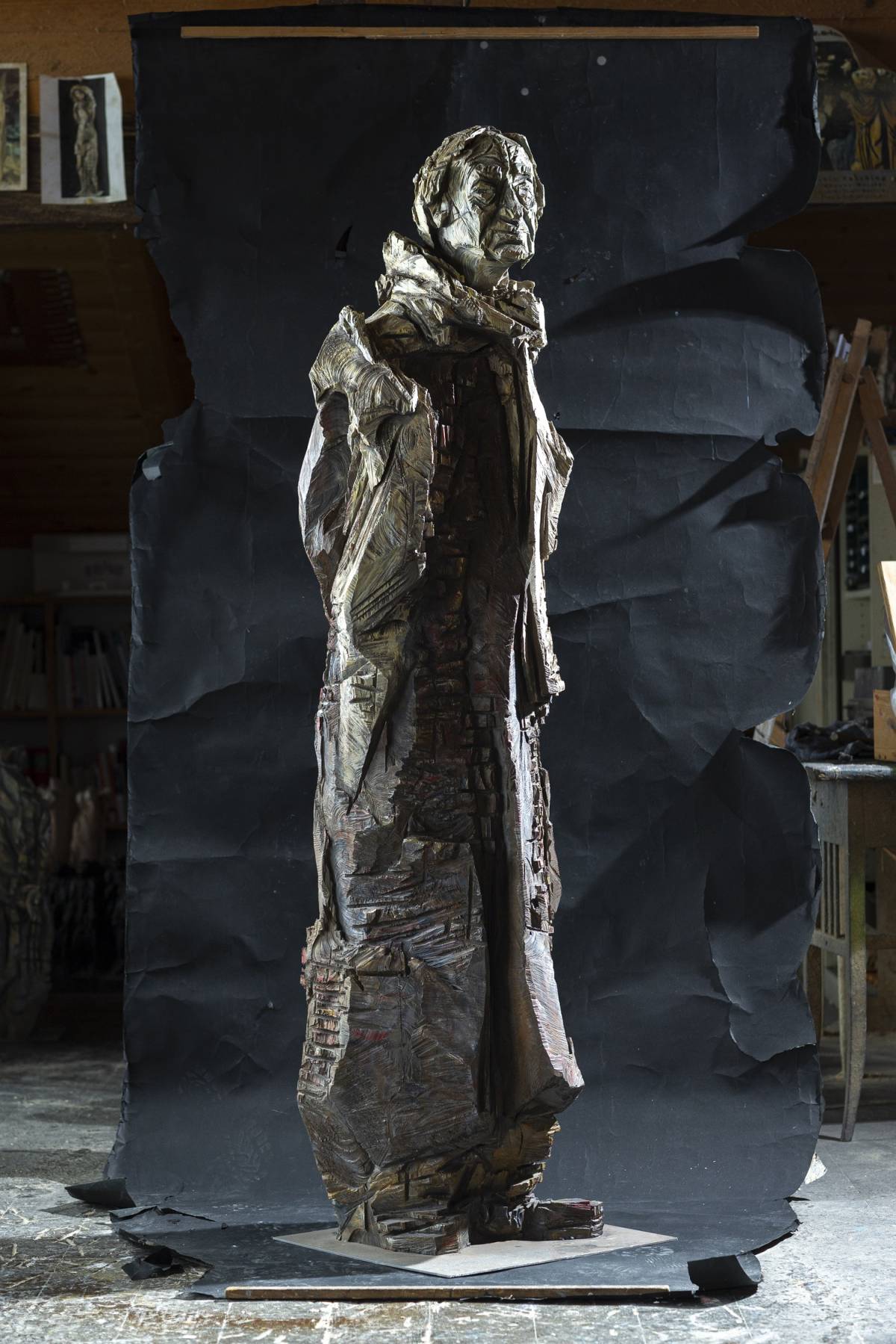 Lebensgroße graue Holzfigur eines Mannes auf Metallplatte mit Schal, sichtbarer Kettensägestruktur und scharfen, tiefen Kanten vor schwarzem Hintergrund