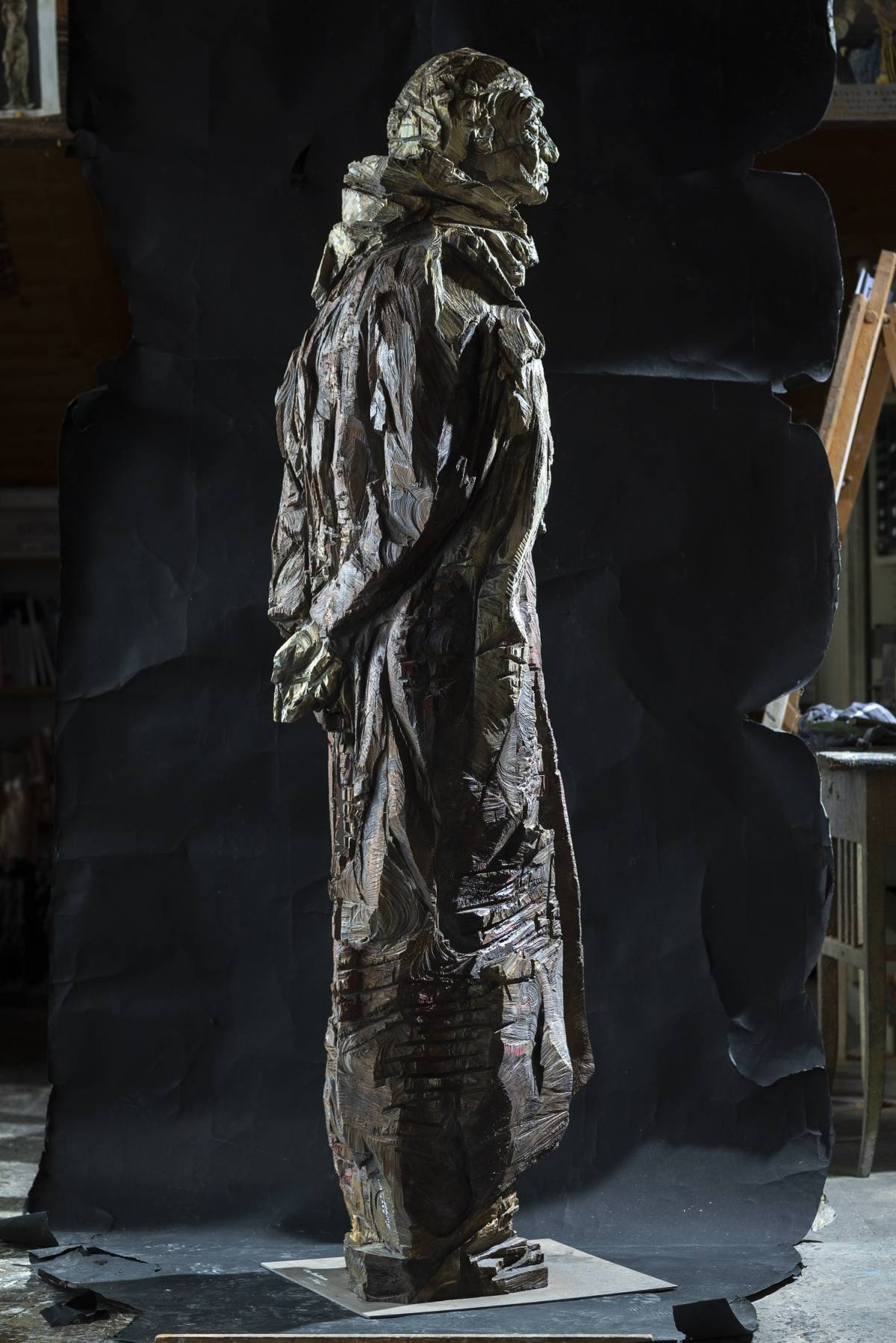 Lebensgroße graue Holzfigur eines Mannes auf Metallplatte mit Schal, von hinten mit überkreutzten Armen, sichtbarer Kettensägestruktur und scharfen, tiefen Kanten vor schwarzem Hintergrund