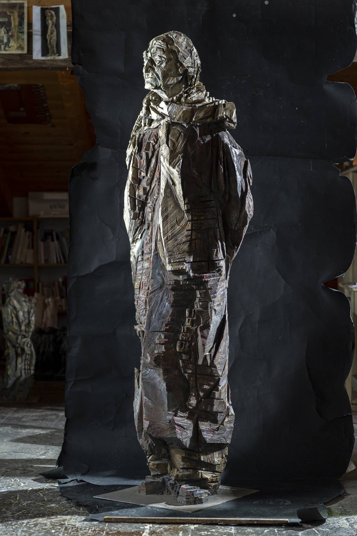 Lebensgroße graue Holzfigur eines Mannes auf Metallplatte mit Schal, vom Betrachter aus nach links schauend, sichtbarer Kettensägestruktur und scharfen, tiefen Kanten vor schwarzem Hintergrund