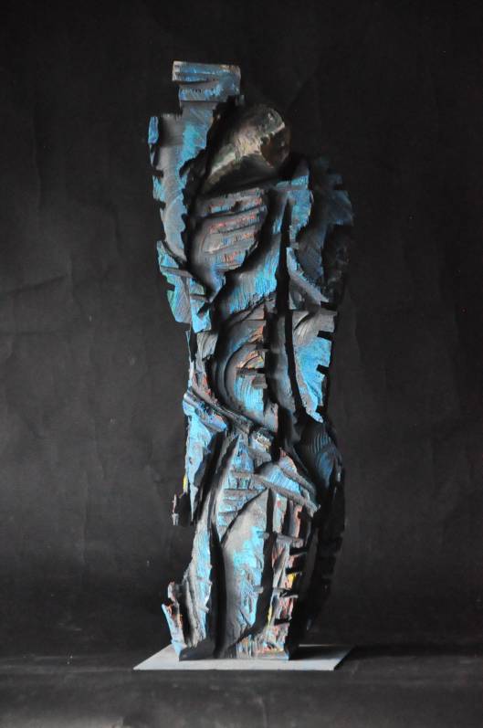 Ganzkörperaufnahme einer blauen Holzskulptur auf Metallplatte mit tiefen, unregelmäßigen Kettensägespuren vor dunklem Hintergrund