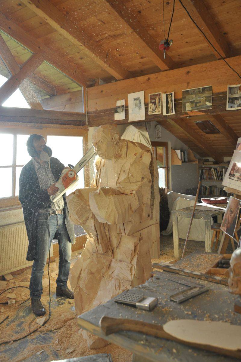 Mann mit Atemmaske und Kettensäge arbeitet in einer Werkstatt an einer lebensgroßen Holzfigur; auf dem Boden liegt Sägemehl.