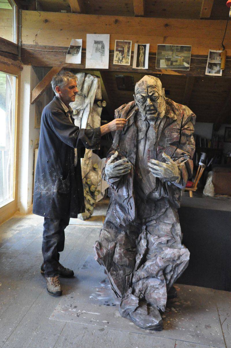 Mann im Arbeitsmantel bemalt in einer Werkstatt mit einem Pinsel eine überlebensgroße, sitzende Holzfigur.