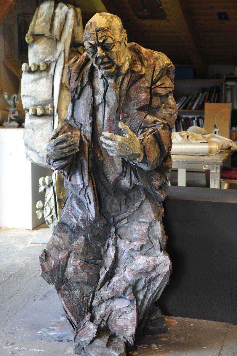 Überlebensgroße, graue, sitzende Holzfigur eines Mannes mit Glatze, der etwas erzählt und dabei mit den Händen gestikuliert.