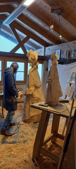 Mann bei der Arbeit mit einer Kettensäge an einer lebensgroßen Holzfigur, rechts davon steht das Modell