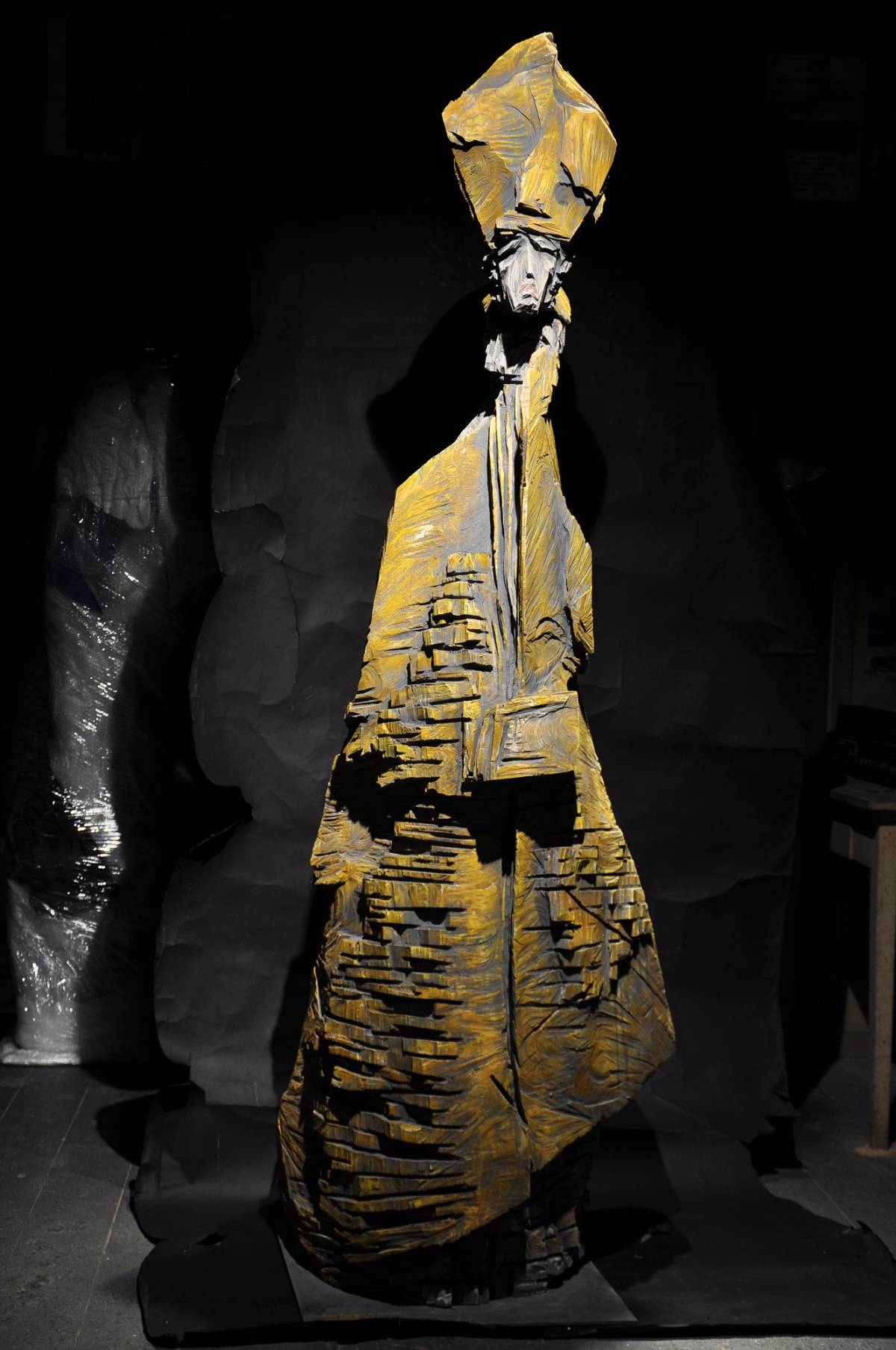 Lebensgroße gelbe Holzfigur mit Bischofsmütze auf Metallplatte vor schwarzem Hintergrund, mit sichtbarer Holzstruktur und unterschiedlichen Kettensägespuren