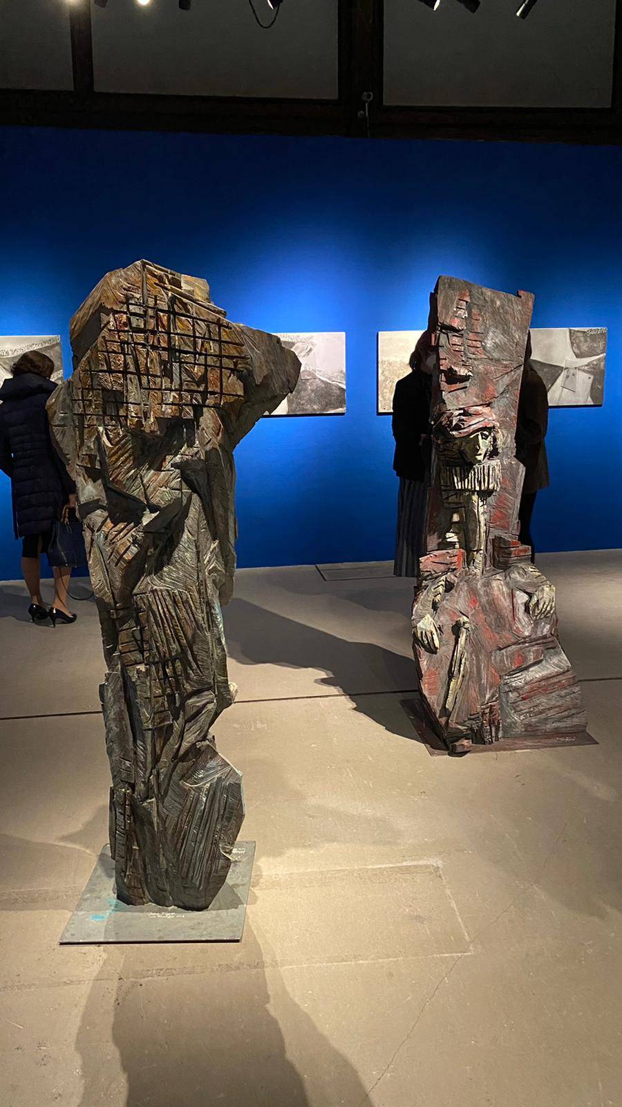 links Rückseite einer Holzfigur mit deutlichen, unregelmäßigen Kettensägespuren, oben bunt und unten in Blau-Grau gestaltet, rechts eine sitzende rote Holzfigur