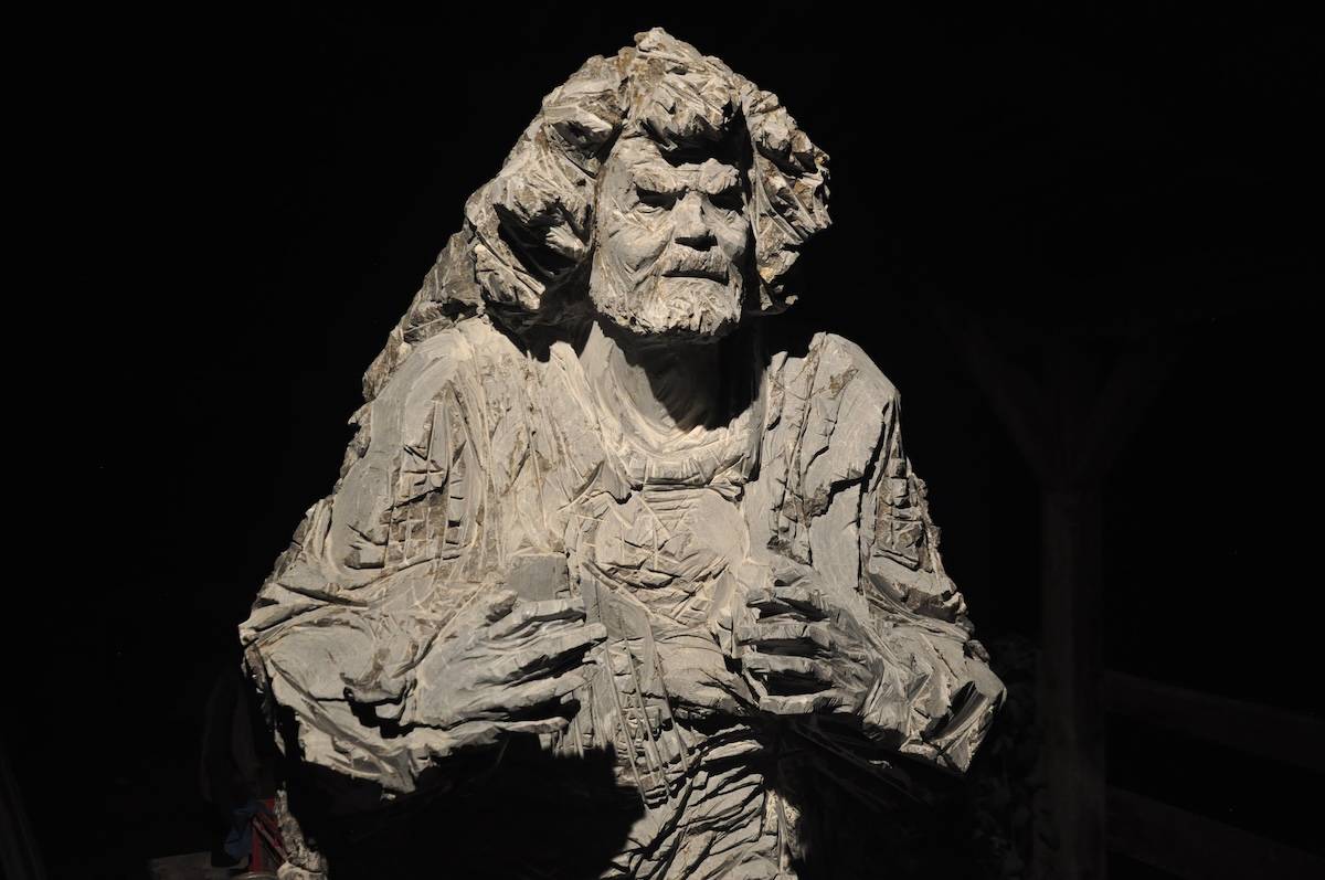 Skulptur aus hellem Stein: Oberkörper und Kopf eines bärtigen Mannes mit ernstem Blick und ausdrucksstarke Händen, kantig herausgearbeitet und vor dunklem Hintergrund beleuchtet